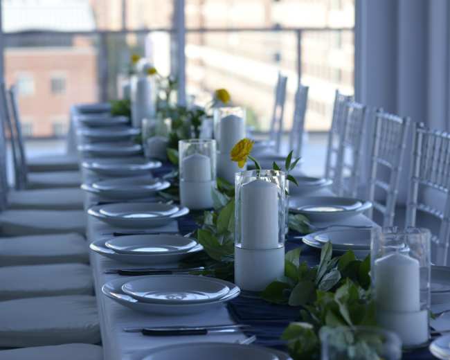 A long banquet table is set with white plates, glass vases containing candles, and greenery, arranged in a bright room with large windows overlooking a cityscape.