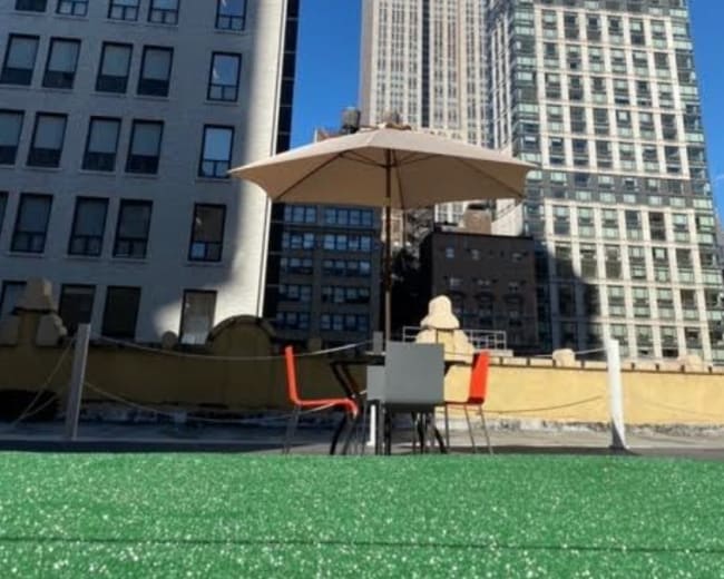 A rooftop space with green synthetic grass features a table and chairs beneath an umbrella, overlooking the Empire State Building.