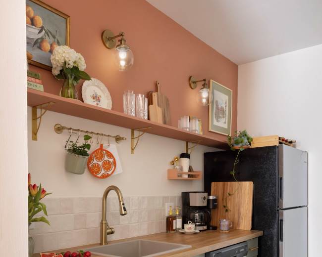 A modern kitchen featuring a double sink with a gold faucet, a coffee maker, a refrigerator, and open shelving displaying kitchenware against a peach-colored wall.