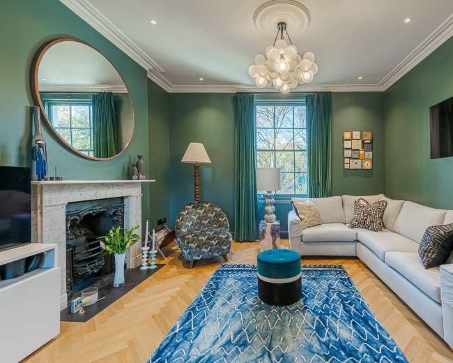 The living room features a white sectional sofa, a blue patterned rug, a large mirror, and a fireplace, complemented by green walls and decorative accents.