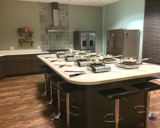 A modern kitchen with an island equipped with several cooking pans, surrounded by high-backed stools and featuring dark cabinets with a patterned backsplash.