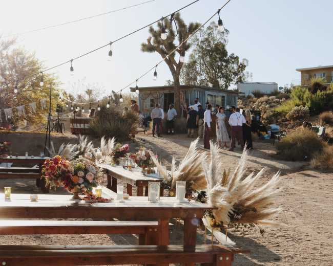 A rustic outdoor wedding setup features long wooden tables with floral arrangements and string lights in a desert setting.