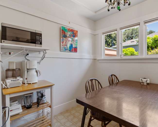A small kitchen features a wooden dining table and chairs, with a microwave and appliances on a shelf, and windows providing natural light.