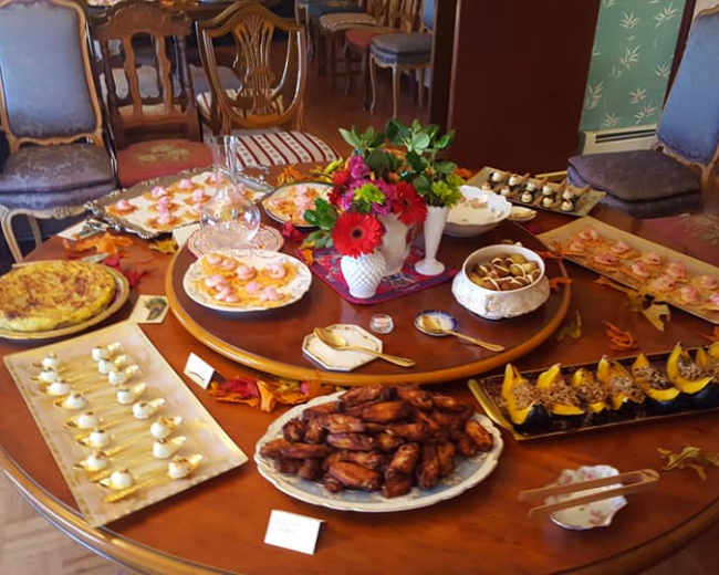 A round wooden table is set with an array of various dishes and desserts, including savory pastries, chicken wings, and beautifully arranged sweets, surrounded by decorated chairs.