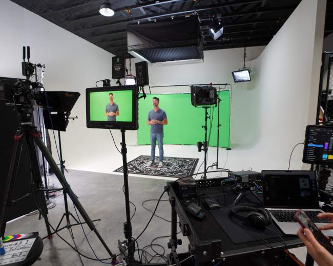 A person stands in front of a green screen, surrounded by video equipment and lighting in a studio setting.