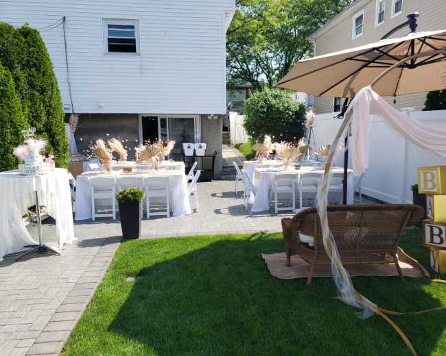 The image shows a backyard setup with tables covered in white cloth, decorated with floral arrangements, chairs, and an umbrella, alongside a green lawn and a cozy seating area.