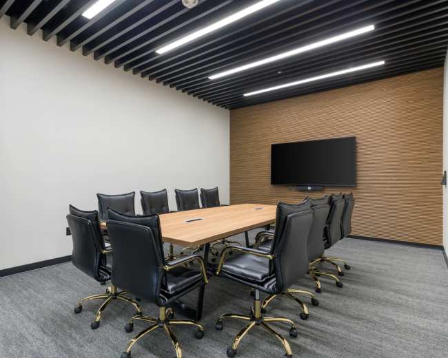 The image shows a modern conference room featuring a large rectangular wooden table surrounded by eight black leather office chairs, with a wall-mounted flat-screen television at one end.