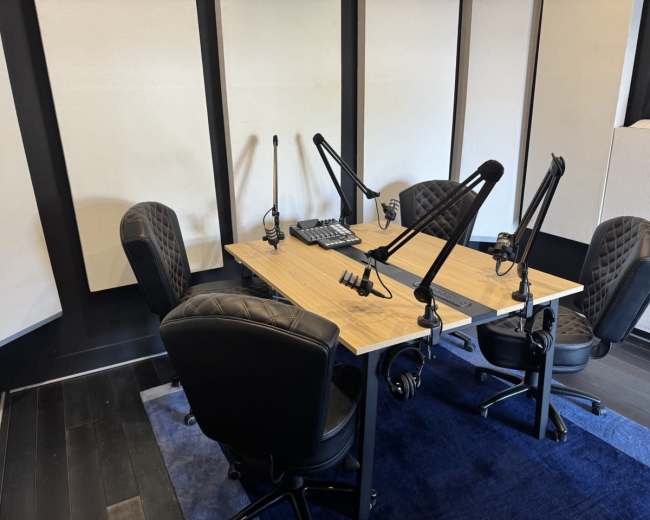 The image shows a podcasting studio with a wooden table, four black rolling chairs, and multiple microphone stands positioned around it.