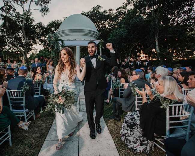 A newlywed couple walks down the aisle, celebrating their marriage as guests applaud and cheer in a garden setting.