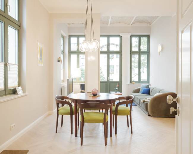 A dining area with a round wooden table surrounded by chairs, set against a backdrop of large green-framed windows and a cozy living space featuring a sofa.