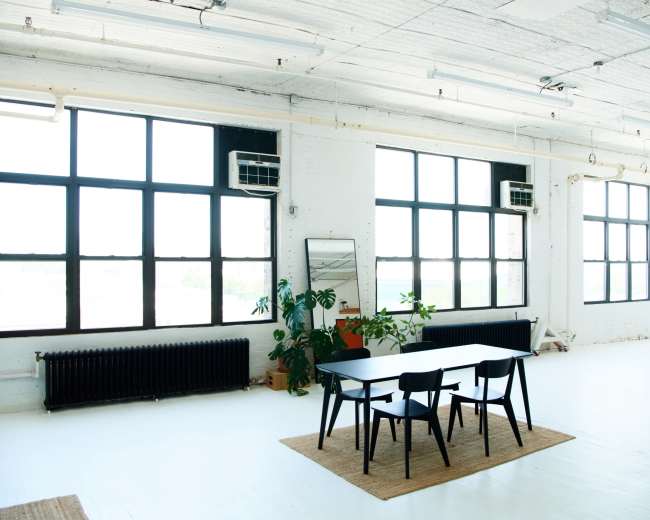 A spacious, well-lit room features a black table with chairs, large windows, a mirror, and potted plants against a white floor.