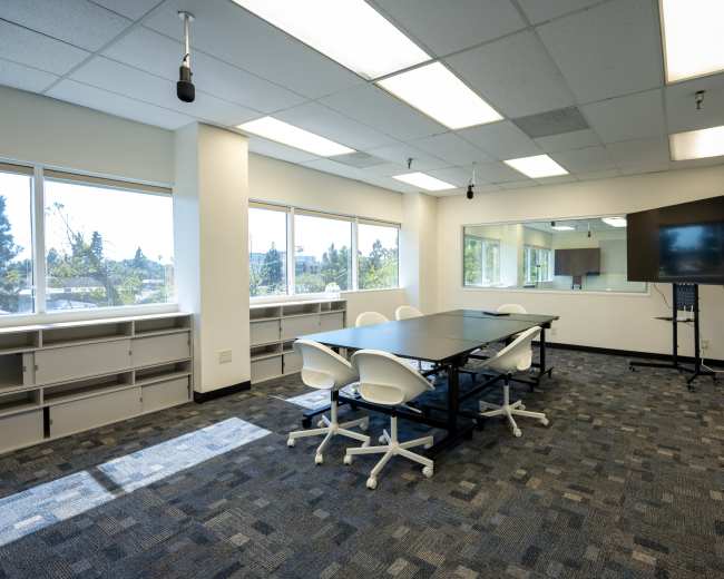 A modern conference room features a large table surrounded by white chairs, with windows providing natural light and a television mounted on the wall.