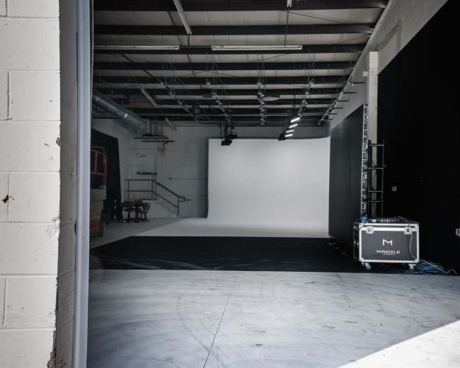 A photography studio with a blank white backdrop, a flat black floor, and equipment stored in the corner.