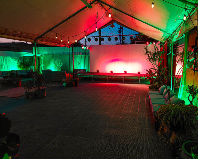 The image shows an indoor space under a tent with colorful lighting, empty seating areas, and potted plants.