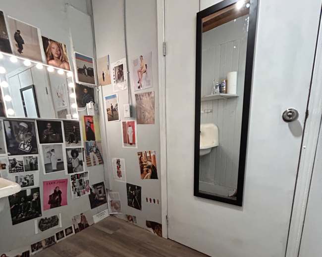The image shows a room with a mirror on the door and walls covered in various photographs, next to a lighted vanity mirror and a small sink.