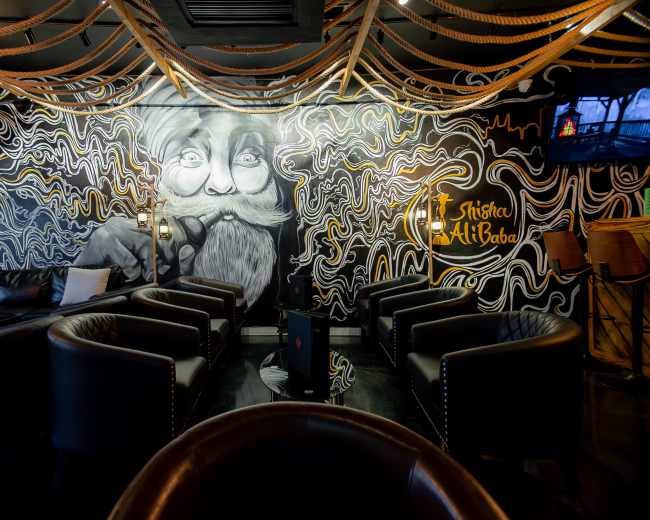 The image shows a stylish lounge area featuring black walls adorned with a large mural of a bearded man, surrounded by decorative rope elements and modern seating arrangements.