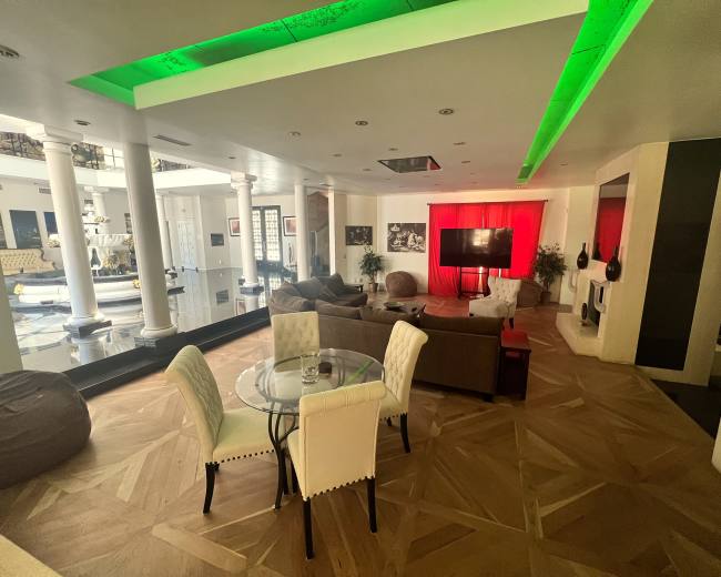 The image shows a spacious living area with modern furniture, including a round glass dining table and a large screen against a red backdrop.