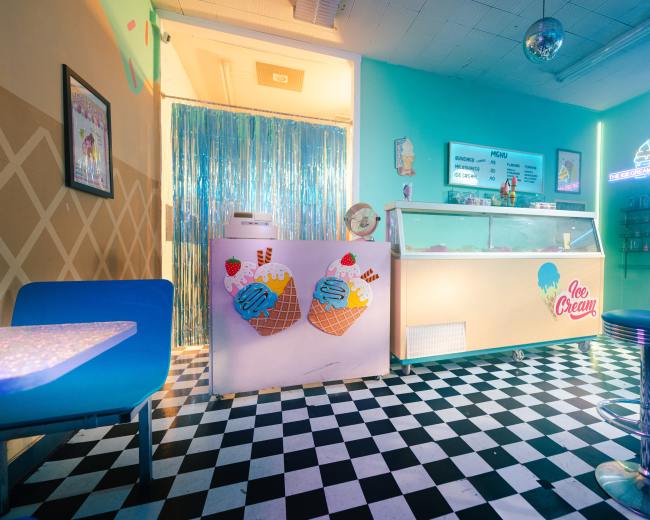 The image shows a brightly lit ice cream shop with a checkered black-and-white floor, a serving counter with an ice cream display, and colorful decor elements.
