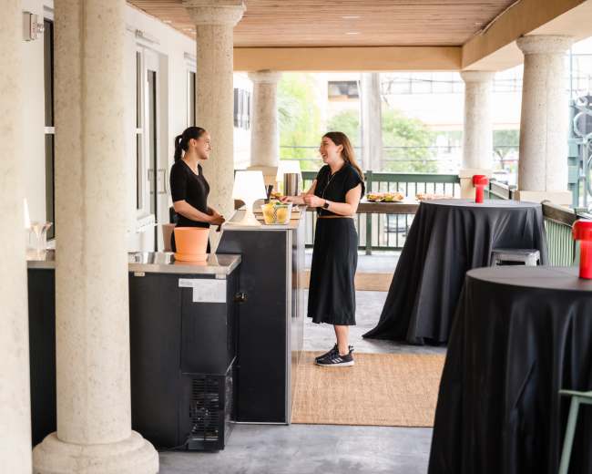 Two women converse at a coffee service station with black tables and food set up in the background.