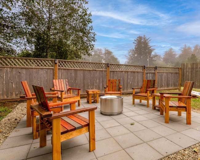 A circular seating area with wooden chairs surrounds a fire pit on a stone patio, enclosed by wooden fencing and trees in the background.