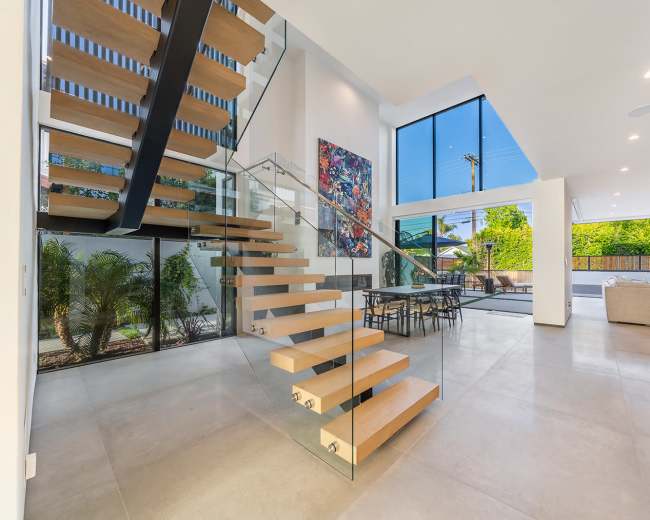 The image shows a modern interior space featuring a floating staircase with wooden steps, large windows providing natural light, and a dining area with a table and chairs.