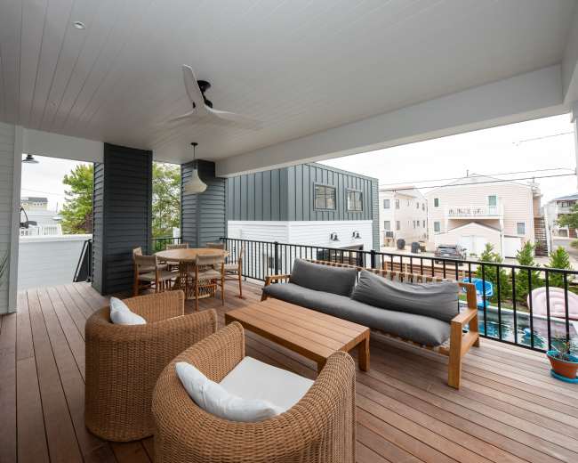 A covered balcony with a wooden floor, featuring a seating area with wicker furniture, a coffee table, and a dining table, surrounded by modern residential buildings.