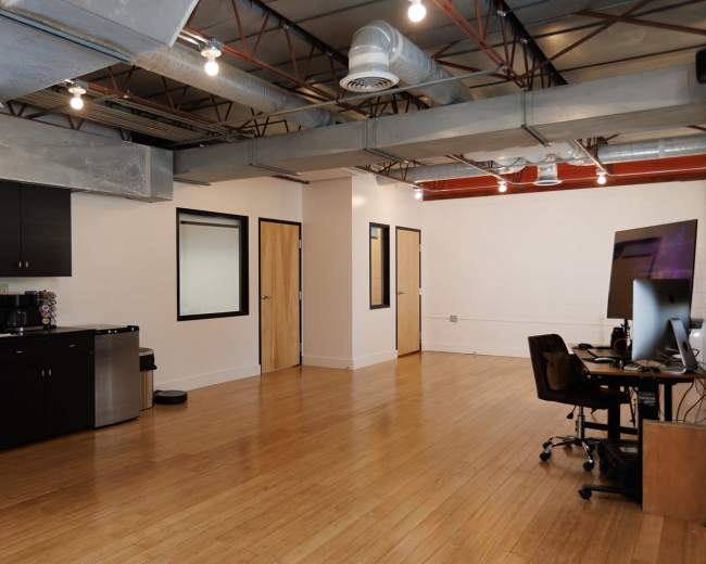 The image shows a modern, open office space with wooden flooring, some kitchen amenities, and a computer workstation against a wall.