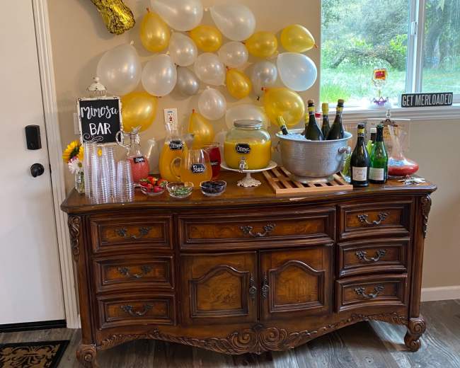 A wooden sideboard displays a variety of drinks, a decorative sign for a mimosa bar, and a backdrop of yellow and white balloons.