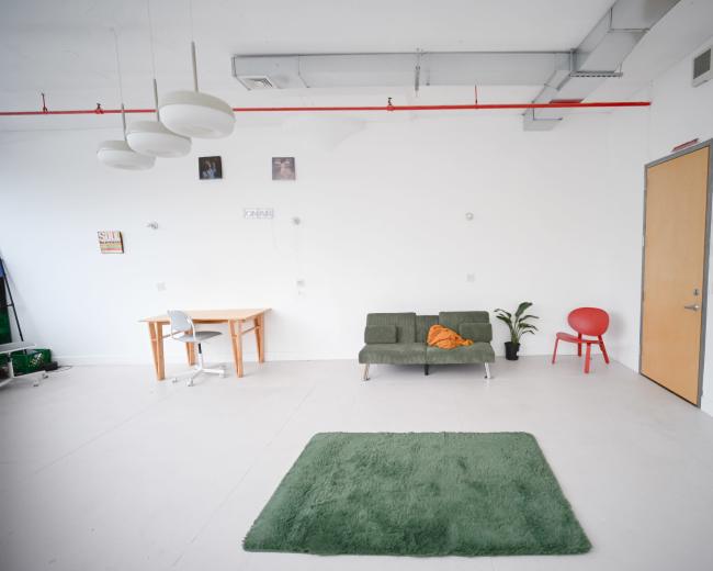 The image shows a minimalist room featuring a green sofa, a small table with a white chair, a red chair, and a green rug on a light-colored floor.