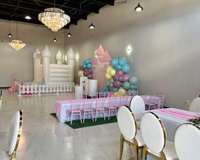 An indoor event space decorated with a balloon arch, a castle-shaped play structure, and tables set with pink and white tablecloths.