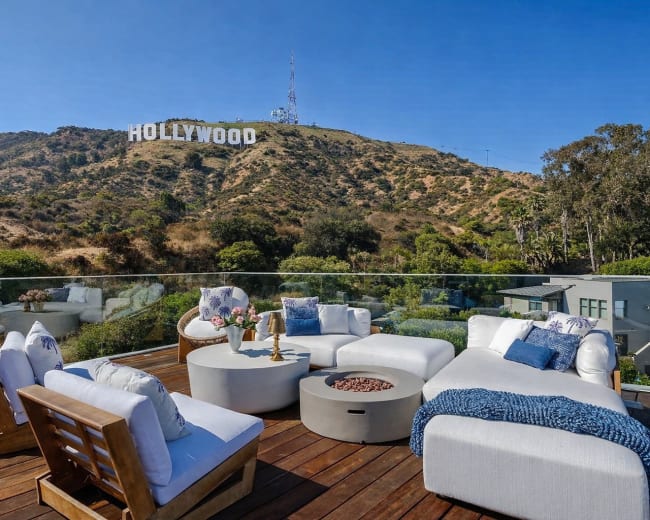 A terrace with modern furniture overlooks the Hollywood sign on a hillside in bright sunshine.