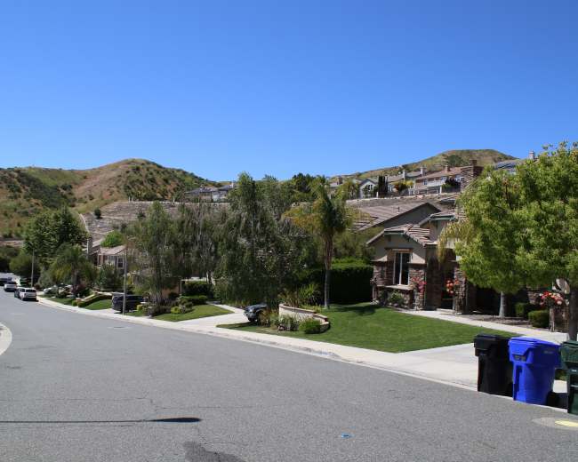 A suburban street curves through a residential neighborhood with houses on either side and hills in the background.