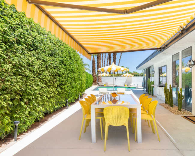 A patio area features a large dining table surrounded by yellow chairs, shaded by a striped yellow awning, with a pool and landscaped greenery in the background.