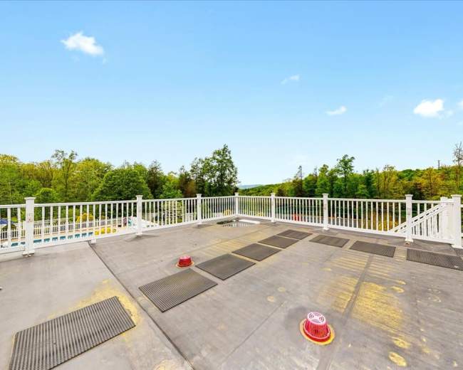 A rooftop deck with a railing overlooking a wooded area, featuring several mats on the surface and a pool visible in the background.