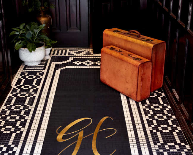 An entryway features a black mat with gold lettering prominently displaying "GE," alongside two vintage orange suitcases and a potted plant.