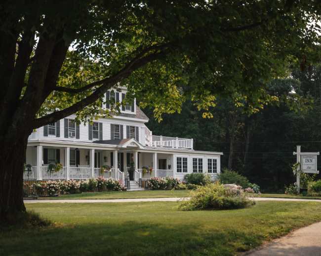 A large, white inn with multiple stories and a wide porch sits under a tree, surrounded by a grassy area and blooming flowers.