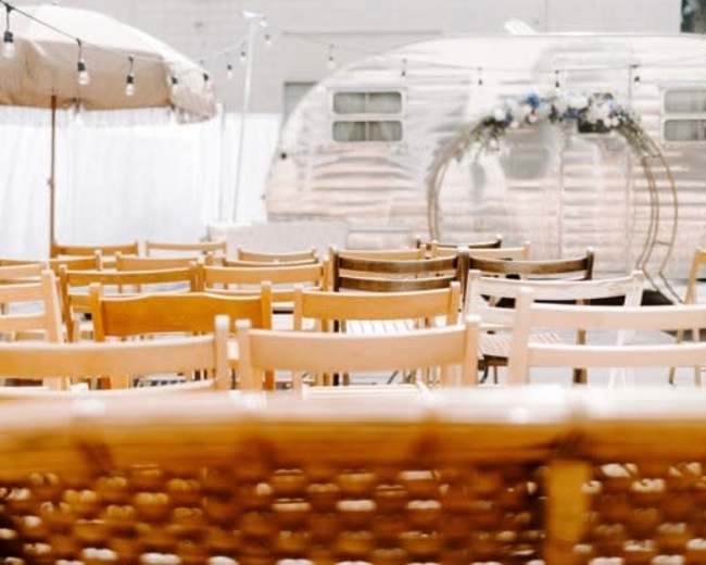 Rows of wooden chairs arranged for an outdoor event, with an Airstream trailer and a shaded area in the background.