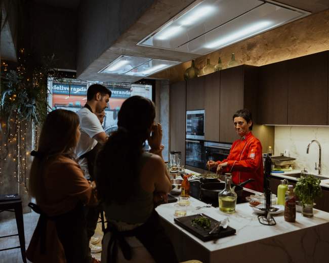 A group of four people watches as a chef in a red coat prepares food in a modern kitchen.