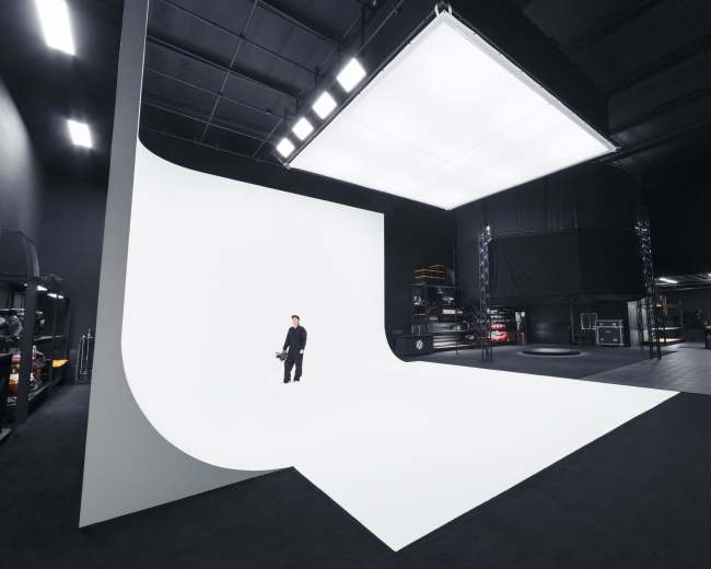 A person stands on a large white backdrop inside a modern studio space with black walls and various equipment visible in the background.