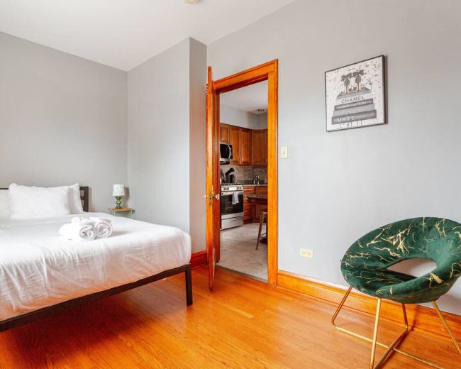 The image shows a simple bedroom with a bed, a green chair, and an open door leading to a kitchen area.