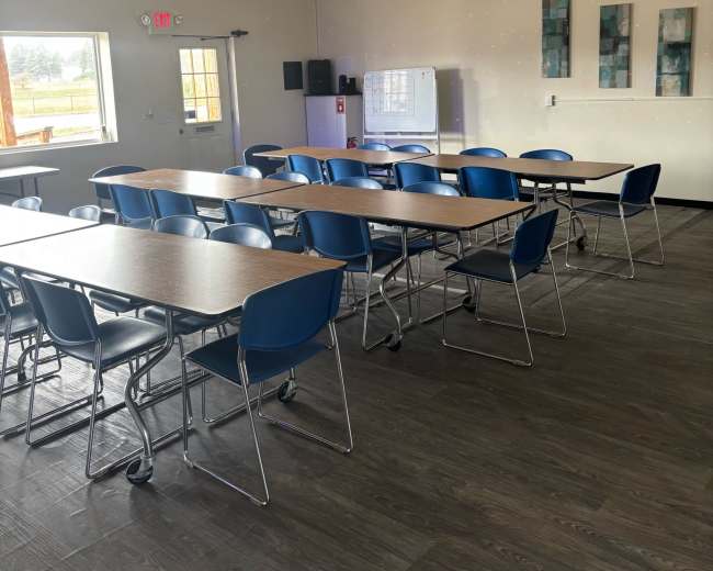 The image shows a spacious classroom with several rows of tables and blue chairs, along with a whiteboard and wall decorations in the background.