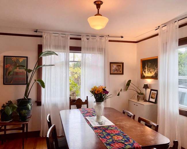 A wooden dining table is set in a light-filled room adorned with plants, floral arrangements, and framed artwork on the walls.