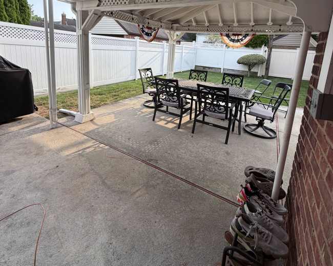 The image shows a covered patio area with a table and chairs, overlooking a lawn with a white fence in the background.