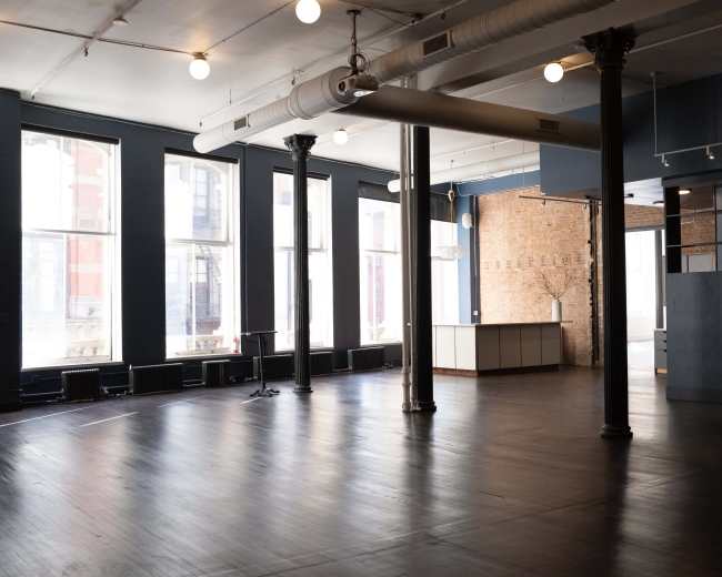 The image shows a spacious, empty room with large windows, wooden floors, exposed brick walls, and columns.