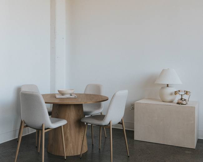 A simple dining area features a round wooden table surrounded by four upholstered chairs, with a lamp and a box on a nearby surface.