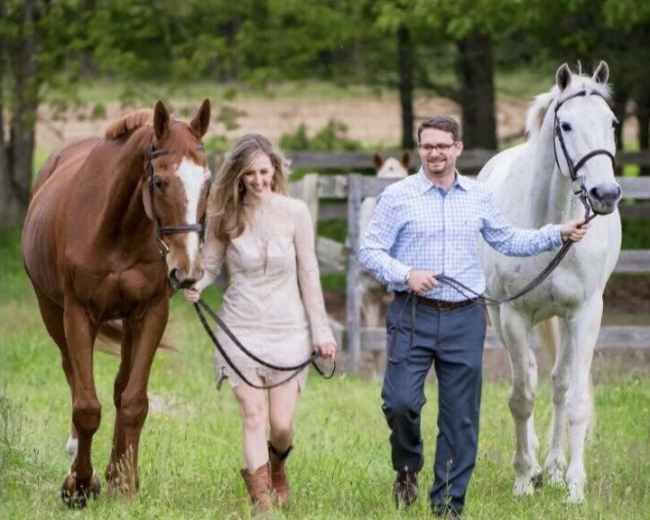 A man and a woman walk two horses along a grassy path near a wooden fence.
