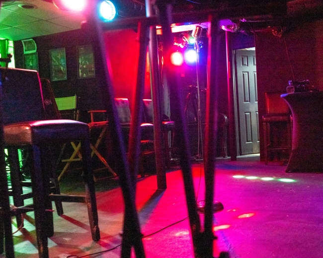 A dimly lit performance space with bar stools, a small stage area, and colorful lights.