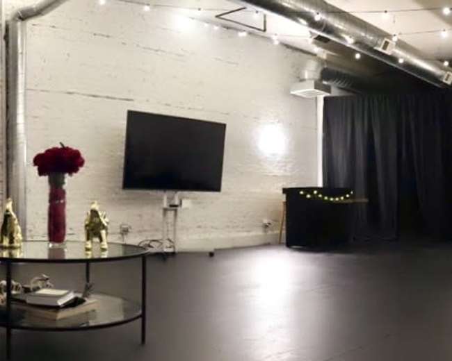 The image shows a spacious studio with a black wooden floor, a TV mounted on the wall, a motorcycle parked in the corner, and string lights overhead.