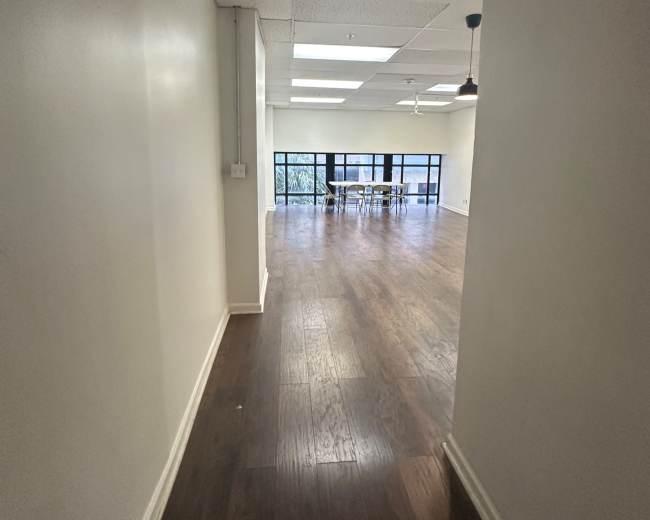The image shows a hallway leading into a spacious room with wooden flooring and large windows at the far end.