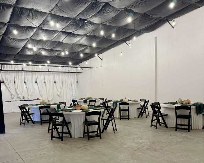 The image shows a spacious indoor event venue with several round tables set with white tablecloths and black chairs, illuminated by overhead lights.